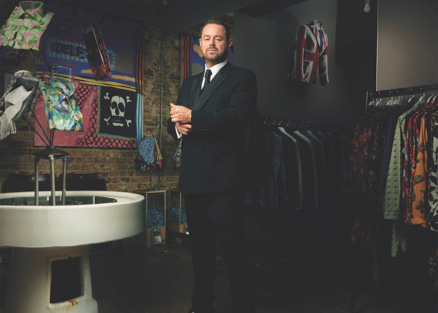 Man in a suit standing in a room with clothing racks and colorful items on the wall.